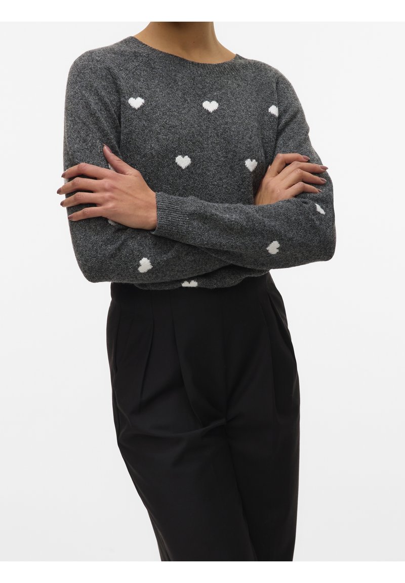 Personne portant un pull gris avec des motifs de cœurs blancs et un pantalon noir taille haute, les bras croisés sur la poitrine.