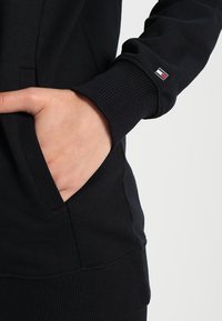 Felpa nera con polsini e orlo a coste. Presenta una tasca laterale e un piccolo patch con logo in rosso, bianco e blu vicino al polsino.