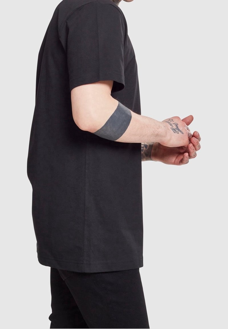 T-shirt en coton noir avec une coupe décontractée, des manches courtes et un ourlet droit. La personne porte un pantalon noir et a un tatouage sur son bras.