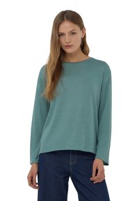 Felpa turchese a maniche lunghe con collo rotondo, texture morbida e vestibilità rilassata. Abbinata a pantaloni in denim blu scuro.