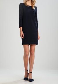 Marinblå stickad klänning med trekvartsärmar, rundad halsringning och rak passform. Matchad med svarta skor med ankelsnöre och rosettdetaljer.