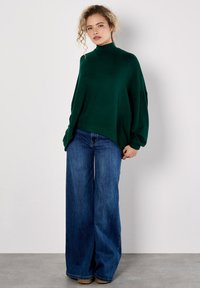 Frau mit lockigem Haar trägt einen dunkelgrünen, übergroßen Rollkragenpullover und weite, blaue Jeans, während sie vor einer schlichten, hellen Wand steht.