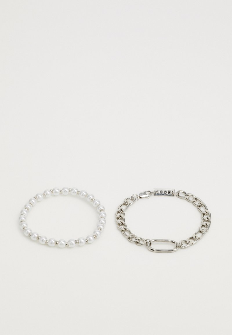 Två armband: ett elastiskt armband i vitt pärldesign och en silverkedja med spänne och ett "ICON"-tagg, med släta ytor och runda former.
