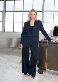 Femme en costume bleu foncé debout pieds nus dans une pièce de style industriel, appuyée sur une table en métal avec un ordinateur portable et un câble orange.