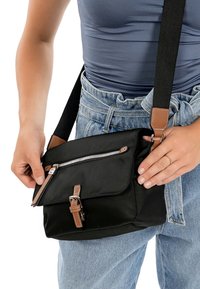 Person trägt blaue Oberbekleidung und Jeans und richtet eine schwarze Umhängetasche mit braunen Lederakzenten und silbernem Reißverschluss.