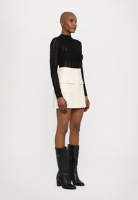 Zwarte doorschijnende top met lange mouwen en gestreept patroon, gecombineerd met een witte minirok met knopen aan de voorkant en zakken, en zwarte kniehoge laarzen.