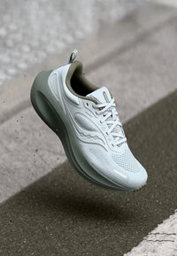 Baskets de course gris clair avec tige en mesh, semelle en caoutchouc et accents verts. Présente un système de laçage et des éléments de design texturés.
