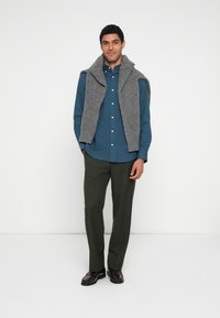 Pilka megzta liemenė ant mėlynos mygtukinės marškinių, derinama su žaliais kelnėmis ir ruda avalyne, neutraliame fone.