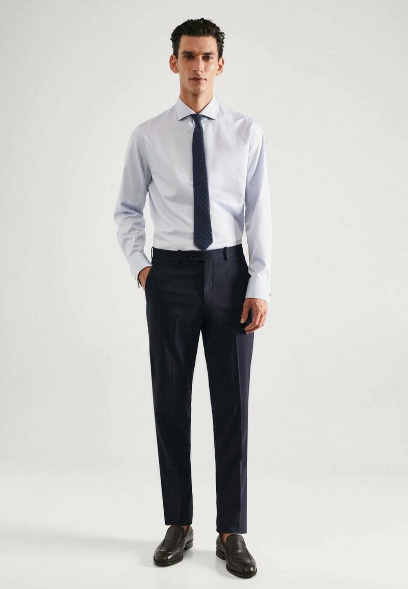 Chemise bleu clair à manches longues, cravate marine à petits pois, pantalon sombre ajusté et chaussures noires à enfiler.