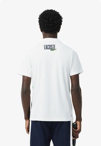 Polo bianco con maniche corte, dettagli verdi e blu navy, con il logo "LACOSTE" e l'emblema del coccodrillo sul retro. Texture liscia.