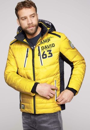 Mann trägt gelb-schwarze wattierte Jacke, öffnet eine Tasche auf der rechten Seite und schaut leicht nach links.