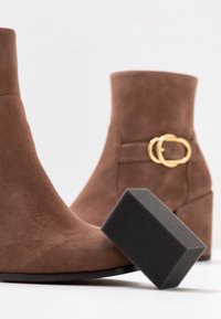 Bottines en suède marron avec un talon bloc, ornées d'une boucle dorée près de la cheville. Une éponge de nettoyage grise est placée devant.