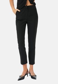 Pantaloni neri su misura con una vestibilità slim, vita media, tasche laterali e pieghe frontali; abbinati a scarpe con tacco a punta nere.