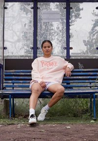 Sudadera Nike rosa claro con el texto "FRANCIA", pantalones cortos blancos con cuadros azules, zapatillas negras y blancas, sentado en un banco azul.