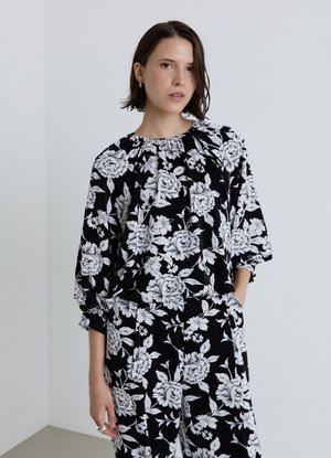 Donna con capelli corti e scuri che indossa un outfit nero con grandi motivi floreali bianchi, una mano in tasca, in piedi davanti a uno sfondo chiaro e uniforme.