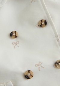 Valge kangas heledate beežide tikanditega lipsumustritel ja läikivate kuldsete nuppudega. Sile tekstuur peene läikega.