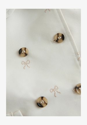 Balts audums ar gaiši bēšiem izšūtiem loku rakstiem un mirdzošām zelta pogām. Gluda tekstūra ar smalku spīdumu.