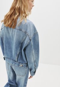 Svetlo modra denim jakna z sproščenim krojem, spuščeni rameni in vidnimi šivi; v kompletu z ujemajočimi se ohlapnimi kavbojkami.