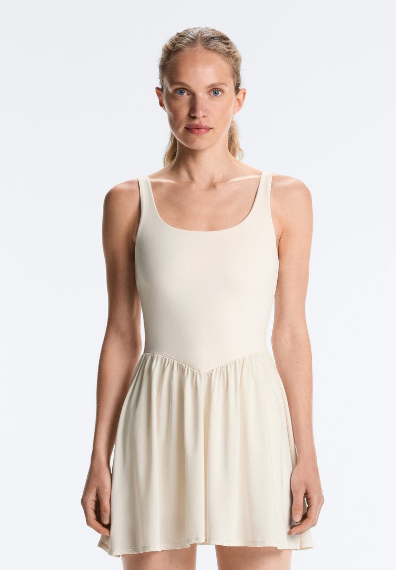 Femme aux cheveux blonds attachés en arrière portant une robe moulante sans manches de couleur crème sur un fond clair uni.