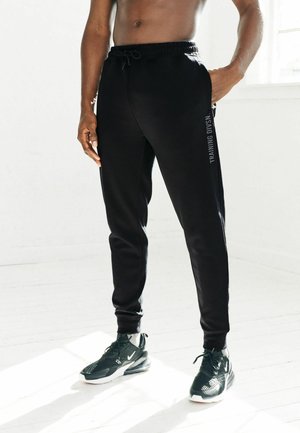 Schwarze Sporthose mit Kordelzug, schmal zulaufendem Design, seitlichen Taschen und "TRAINING DIVSN" vertikal auf dem Bein bedruckt.