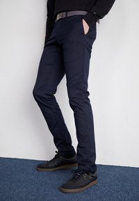 Pantalon en coton marine à coupe slim, avec poches latérales et une ceinture grise. Porté avec des baskets noires sur une surface de tapis bleu.