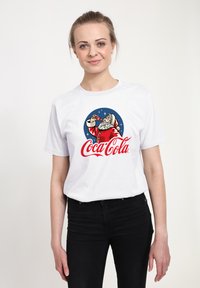 Bílý bavlněný tričko s kulatým logem Santa Clause, který pije z lahve Coca-Coly, s červenou a modrou barvou doplněnou bílými hvězdami.
