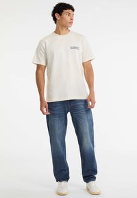 Młody mężczyzna w kremowej koszulce Guess Jeans, niebieskich dżinsach i białych trampkach stoi na tle białego tła.