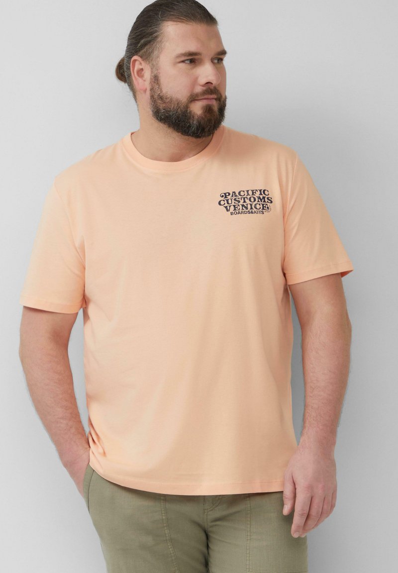 Pfirsichfarbene Baumwoll-T-Shirt mit kurzen Ärmeln und Rundhalsausschnitt. Auf der Vorderseite befindet sich schwarzer Grafiktext mit der Aufschrift "Pacific Customs Venice Boardski."