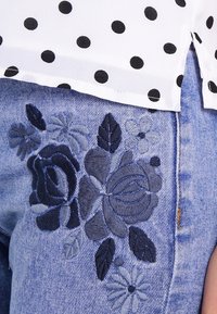 Dżinsy z ciemnoniebieską kwiatową haftem na udzie, zestawione z białą bluzką w czarne kropki.