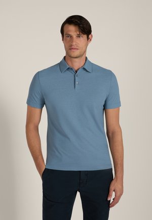 Uomo in piedi con le mani in tasca, indossa una polo azzurro chiaro a maniche corte e pantaloni scuri su uno sfondo semplice.