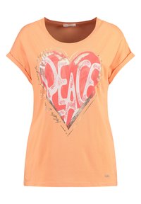 Camiseta de manga corta color melocotón, hecha de algodón suave, con un gran gráfico de corazón rojo que presenta la palabra "PAZ" en blanco, acentuada con detalles metálicos.