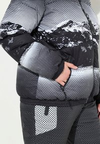 Chaqueta acolchada de patrones en blanco y negro con un bolsillo con cremallera, que presenta una textura esponjosa y un diseño en medio tono. La chaqueta tiene una silueta ajustada.