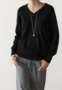 Pull en tricot noir avec un décolleté en V, manches longues et coupe décontractée, associé à un pantalon en denim gris clair. Collier avec pendentif en argent visible.