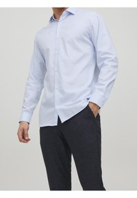 Muž v bledě modré košili s dlouhým rukávem a tmavých kalhotách, s jednou rukou v kapse, na jednobarevném pozadí.