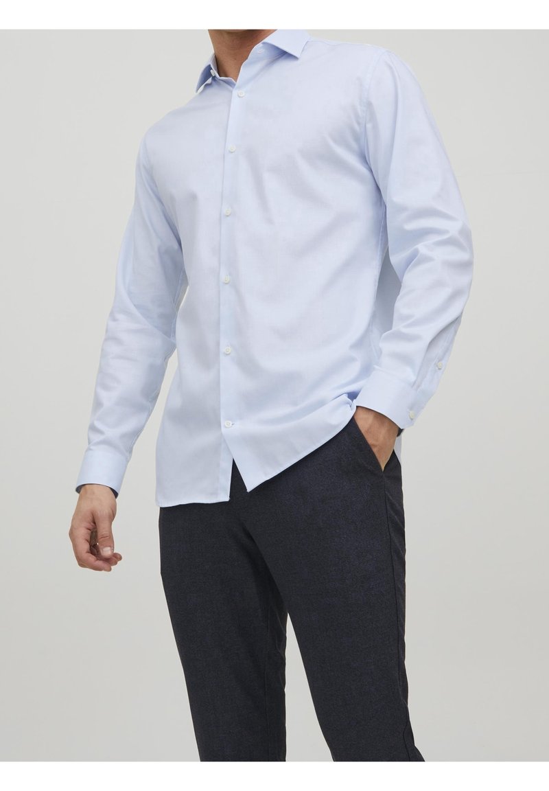 Muž v bledě modré košili s dlouhým rukávem a tmavých kalhotách, s jednou rukou v kapse, na jednobarevném pozadí.