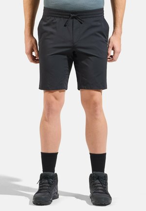 Mann trägt schwarze Shorts mit Kordelzug, schwarze Sportsocken und schwarze Sportschuhe, steht vor hellem Hintergrund.