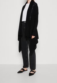 Svart oversized cardigan med vertikal ribbad textur, öppen fram, och långa ärmar, kombinerad med svarta raka jeans och platta svarta skor.