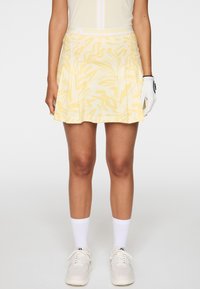 Femme portant une jupe plissée jaune à motifs, des chaussettes blanches, des baskets blanches et un gant de golf blanc à la main droite.