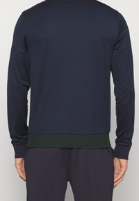 Marinblå långärmad sweatshirt med grön ribbad nederkant, mjuk textur, figurnära design och avslappnad silhuett, visas från baksidan.