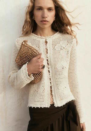 Woman wearing a cream crochet cardigan and dark skirt, holding a woven clutch purse with loose blonde hair against a light background.