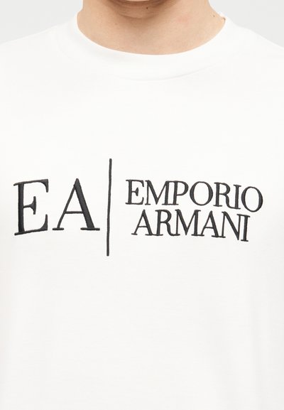 Λευκό μπλουζάκι με στρογγυλή λαιμόκοψη και μαύρο κεντημένο λογότυπο "EA | Emporio Armani" στο στήθος, φορεμένο από άτομο που φαίνεται από το λαιμό και κάτω.