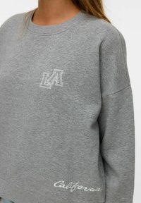 Felpa grigia con scollo rotondo, vestibilità over size, con accenti ricamati in bianco "LA" e "California". Tessuto morbido, design casual.