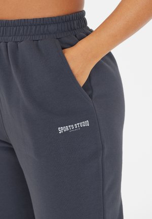 Dunkelblaue Sweatpants mit elastischem Bund, ausgestattet mit einer Seitentasche und einem weißen, aufgestickten "SPORTS STUDIO"-Logo in der Hüfthöhe.