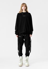 Sudadera negra de gran tamaño con un bolsillo frontal; combinada con pantalones jogger negros que presentan acentos negros en forma de alas y detalles de etiqueta blanca en contraste.