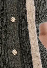 Πλεκτό κατ' αγκαλιά σε σκούρο πράσινο χρώμα με χοντρή υφή, διαθέτοντας διακοσμητικά κουμπιά σε χρυσό τόνο και μια αντίθετη μπεζ λωρίδα κατά μήκος της άκρης.