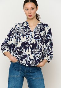 Marineblaue Bluse mit weißem Blumenmuster, ausgestattet mit einem V-Ausschnitt, lockerem Schnitt und dreiviertellangen Ärmeln. Das Material wirkt leicht und fließend.