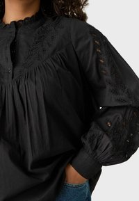 Blusa nera in cotone con collo alto, busto arricciato e dettagli in pizzo intricati sulle maniche. Tessuto testurizzato con maniche voluminose.