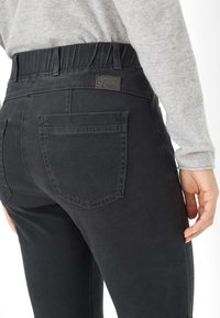 Pantaloni in denim nero con una cintura elastica, dotati di due tasche posteriori e un'etichetta in tessuto testurizzato, ripresi da una vista laterale.