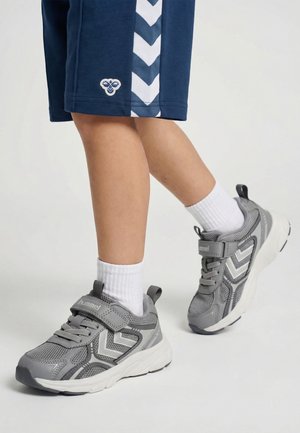 Kind draagt grijze en witte sneakers met klittenbandsluitingen, witte sokken en marineblauwe korte broek met witte chevronstrepen.