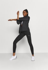 Giacca sportiva nera con polsini elasticizzati, abbinata a leggings neri a vita alta con logo Puma bianco. Sneakers bianche con dettagli.